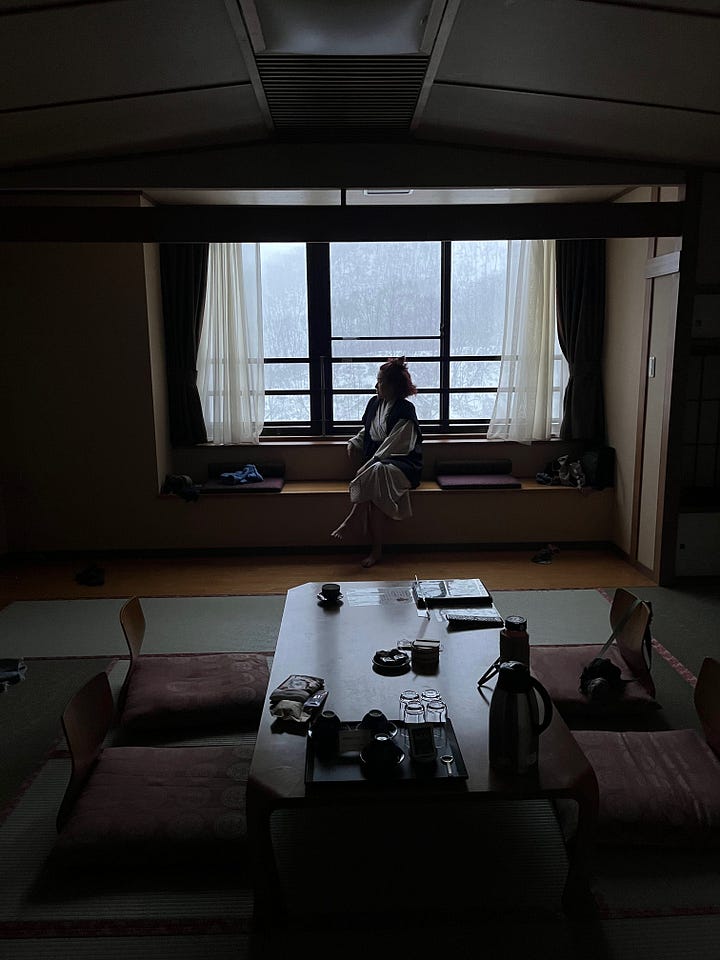 1. Myself in a white rain jacket with a neon blue hat that has Yu & Me embroidered on it next to a large stuffed teddy bear. 2. A brown bear lying on its back with one hand holding its foot and the other hand out to catch food. 3. Lake Kuttara same as the first image 4. View of the hotel room with the window showing the snow outside while a person is sitting looking out the window backlit wearing a traditional yukata robe. 5. A charcoal grill lit and ready with Wagyu beef, marinated pork, small fishes, and a semi raw egg on plates before the grill. 