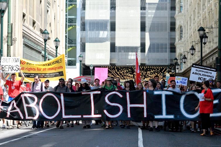 Crowd of people walk down a city street holding a large black banner that says 'Abolish ICE' Crowd of people walk down a city street holding a large black banner that says 'Abolish ICE'