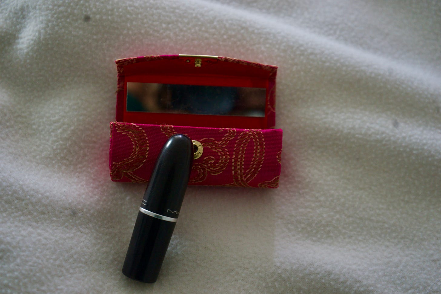 A black lipstick stands upright before a small red patterned case with a partially revealed mirror, centered against soft white fabric, composed with restrained framing and intimate depth to suggest ritual and calibration. A black lipstick stands upright before a small red patterned case with a partially revealed mirror, centered against soft white fabric, composed with restrained framing and intimate depth to suggest ritual and calibration.