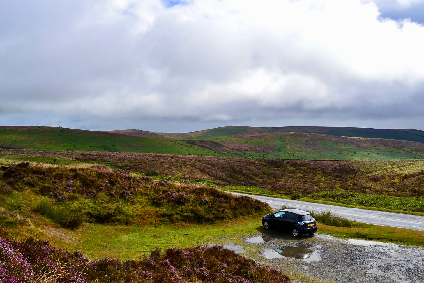 A quiet landscape of the Yorkshire moors at dawn, seen from inside a car, with soft golden light stretching across open hills, evoking stillness, reflection, and the feeling of waiting in silence.