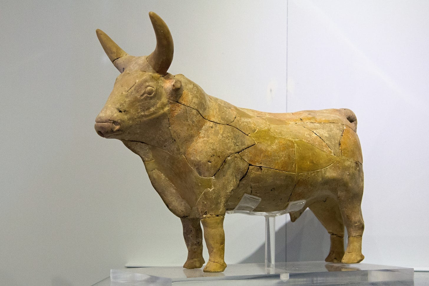 Ceramic figurine of a horned bull. The figurine is heavily cracked and has been glued back together. The whole thing is a putty or yellowish-tan color. The animal has a large head, heavy neck and shoulders with a large dewlap flapping from chin to chest. The genitalia is also visible.