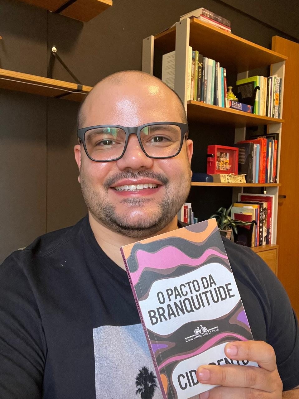 Homem branco, de óculo segura um livro, em sua biblioteca pessoal