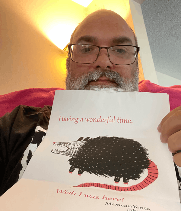 A photo of me holding a letter with a picture of a opossum along with text that reads, “Having a wonderful time, wish I was here.” Signed MexicanYenta