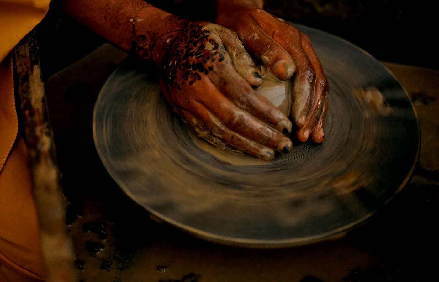 A close-up photograph of a pair of hands, decorated with henna, molding wet clay on a spinning pottery wheel — symbolizing the creative process as the union of intention, resistance, and form.