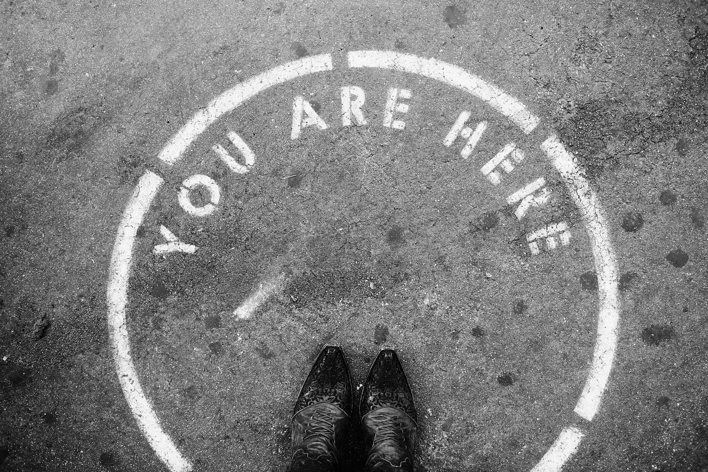 black and white photo of two feet, in cowboy boots, standing in a circle with the words You are Here. 