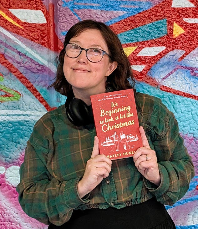 Hayley is holding a printed copy of her book against a backgrop of colourful graffiti