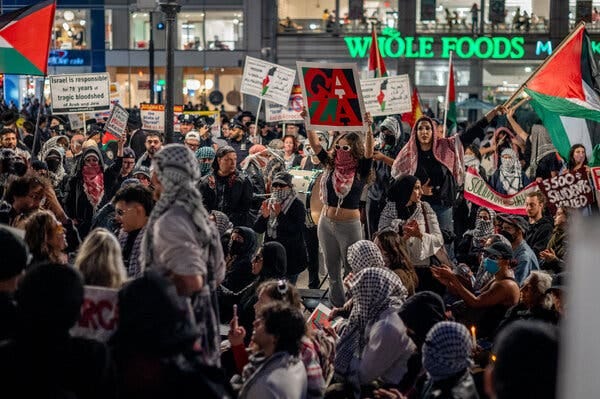 A crowd of protesters, some wearing kaffiyehs and holding signs and Palestinian flags, stand in a plaza at night. A crowd of protesters, some wearing kaffiyehs and holding signs and Palestinian flags, stand in a plaza at night.