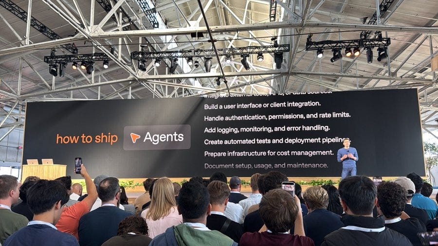 A large screen displaying text about agent development, including "how to ship" and "Agents" with bullet points on building user interfaces, authentication, logging, testing, deployment pipelines, and documentation setup. A person stands on stage speaking to an audience, with people seated and some holding phones. The setting is an indoor venue with a high ceiling, visible beams, and stage lighting. A large screen displaying text about agent development, including "how to ship" and "Agents" with bullet points on building user interfaces, authentication, logging, testing, deployment pipelines, and documentation setup. A person stands on stage speaking to an audience, with people seated and some holding phones. The setting is an indoor venue with a high ceiling, visible beams, and stage lighting.