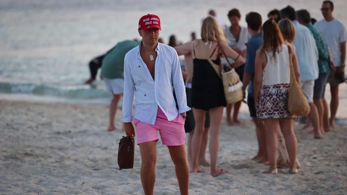 Un uomo cammina su una spiaggia affollata al crepuscolo, indossando un cappellino rosso con la scritta bianca "MAKE AMERICA GREAT AGAIN". Indossa una camicia azzurra leggera sbottonata che rivela una collana con un ciondolo a forma di lettera "B", pantaloncini rosa acceso e tiene in mano una piccola borsa scura in pelle. Sullo sfondo sfocato si vedono diverse persone in abbigliamento estivo che chiacchierano e camminano lungo la riva, vicino all'acqua. Un uomo cammina su una spiaggia affollata al crepuscolo, indossando un cappellino rosso con la scritta bianca "MAKE AMERICA GREAT AGAIN". Indossa una camicia azzurra leggera sbottonata che rivela una collana con un ciondolo a forma di lettera "B", pantaloncini rosa acceso e tiene in mano una piccola borsa scura in pelle. Sullo sfondo sfocato si vedono diverse persone in abbigliamento estivo che chiacchierano e camminano lungo la riva, vicino all'acqua.