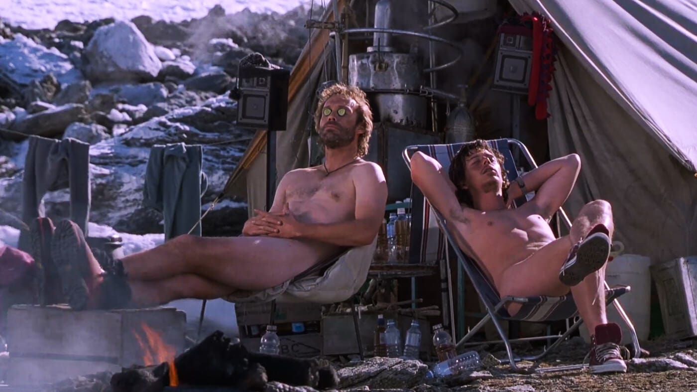 Two nude male climbers sunbathe in camping chairs beside a fire outside their base camp tent, surrounded by snow and outdoor gear.