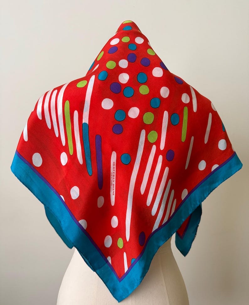 May include: A red silk scarf with a vibrant pattern of white, green, blue, and purple dots and lines. The scarf has a bright blue border and the text "GEOFFREY BEENE" is visible. The scarf is displayed on a mannequin. May include: A red silk scarf with a vibrant pattern of white, green, blue, and purple dots and lines. The scarf has a bright blue border and the text "GEOFFREY BEENE" is visible. The scarf is displayed on a mannequin.