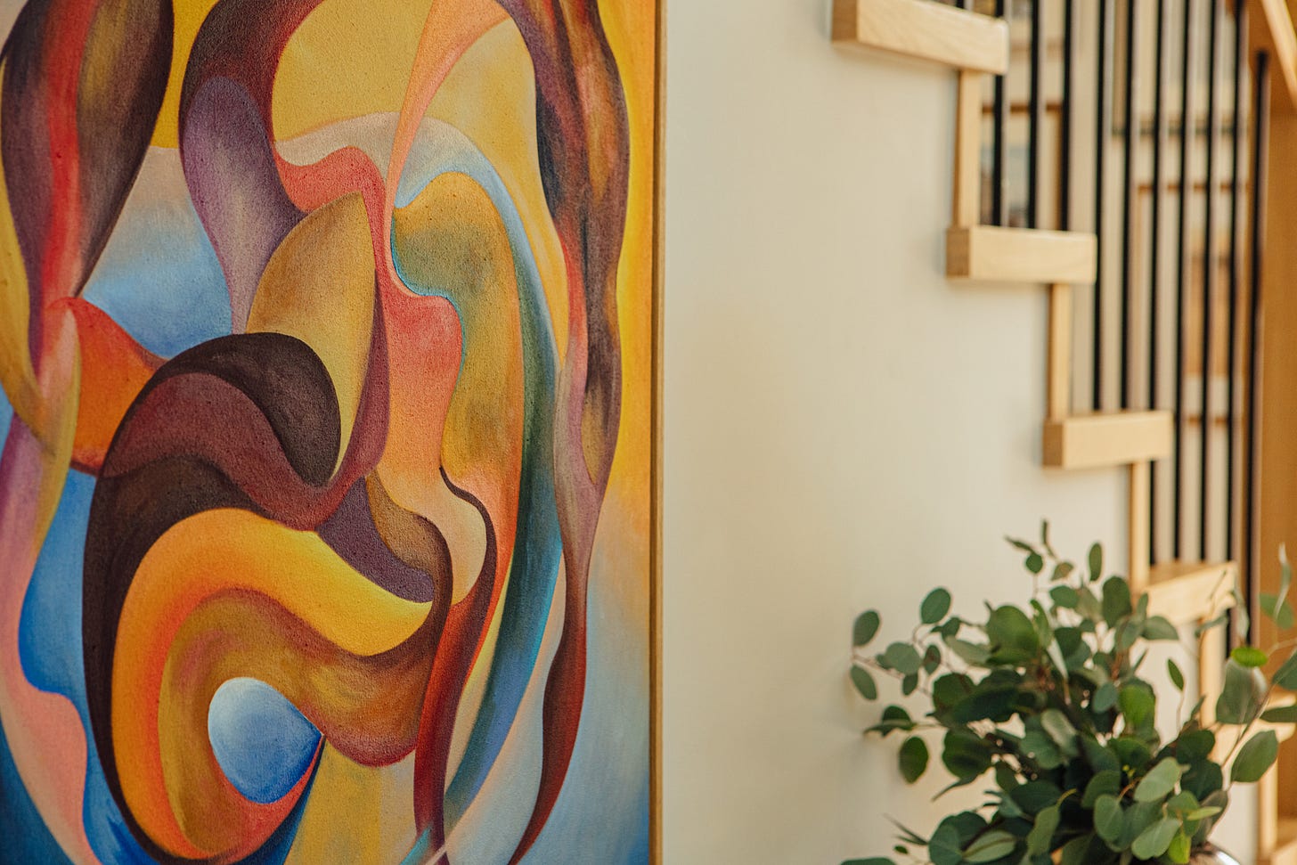Close-up of an abstract painting by Alex Maceda installed in a contemporary home. The composition features layered organic shapes in warm yellow, orange, burgundy, teal, and blue within a light oak frame. Part of an oak staircase and a vase with greenery are visible, showing how the artwork sits within the architecture of the space.