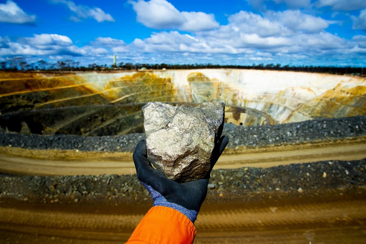 A gloved hand holding a nickel ore rock with an open-cut mine in the background