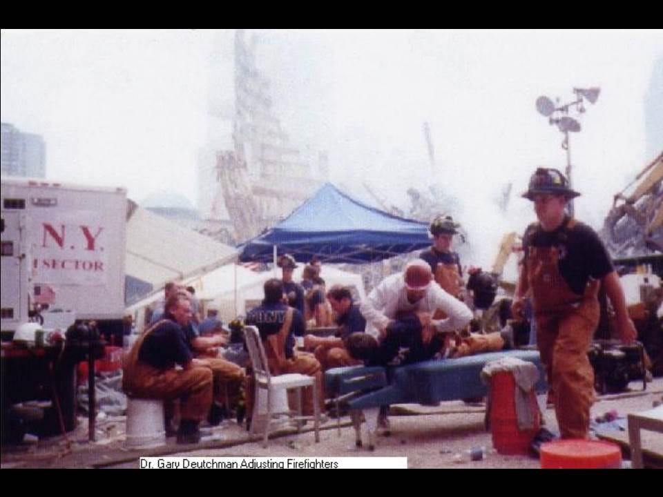 Firefighters in turnout gear rest under a blue canopy while Dr. Gary Deutchman adjusts one on a table, rubble behind