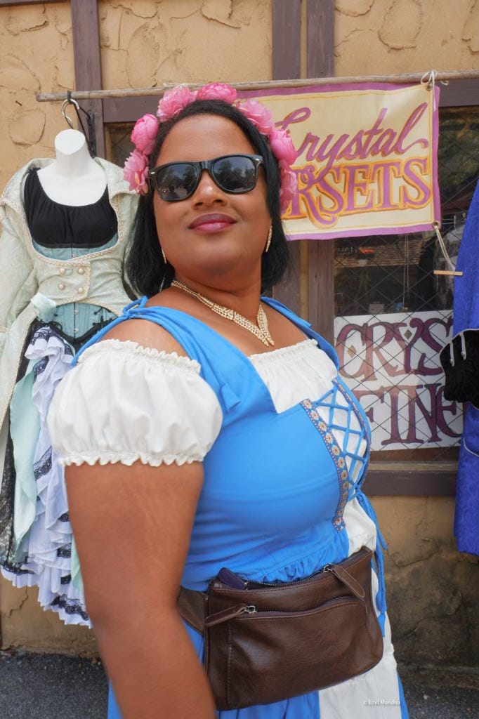 The author wearing a blue Renaissance Faire dress and sunglasses in front of a corset shop.