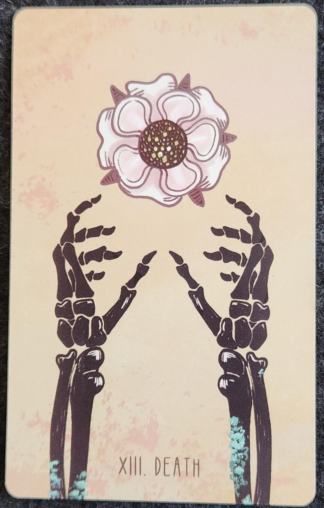 A tarot card labelled XIII. Death. Two black skeletal hands reach up from the bottom of the card, reaching for a white and light pink flower. The centre of the flower is speckled with white dots, looking a bit like a starry sky. The arms of the skeleton have green moss growing on them. The background of the card is a light beige.