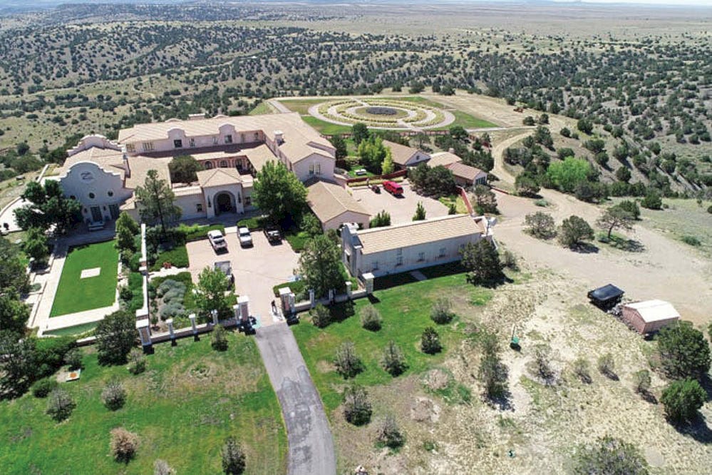 A mansion with several parked cars and a person at the gate, and more distantly outbuildings and gardens A mansion with several parked cars and a person at the gate, and more distantly outbuildings and gardens