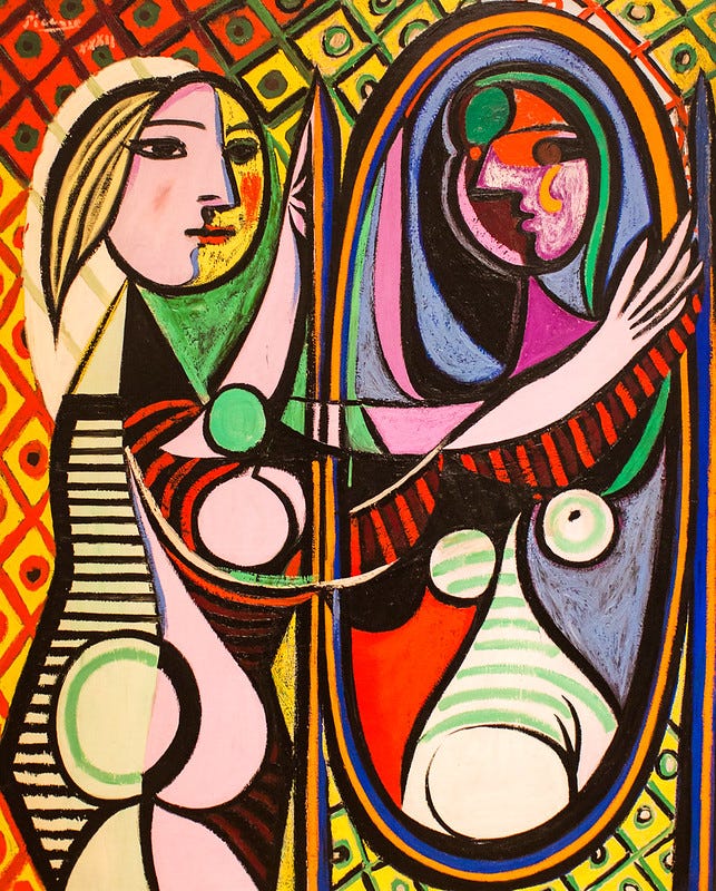 "Girl Before a Mirror" by Pablo Picasso. The painting uses Picasso’s signature Cubist style, breaking the woman’s form into fragmented, geometric shapes. The woman is shown in profile on the left side, facing the mirror on the right. Her skin is depicted in bright yellow and pink. Her hair is in wavy, curved lines, with blues and purples. The reflection in the mirror is more distorted and dark. The reflection shows a fragmented, angular version of the woman, with large, mismatched eyes and a face in harsh shades of green and red. The background is filled with vibrant, geometric patterns, including diamond shapes and vivid colour blocks in purple, green, yellow, and red.