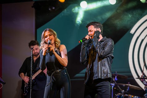 Alina Crișan & Keo doi artiști cântă împreună pe scenă, față în față, fiecare ținând câte un microfon. Artista poartă o ținută neagră, iar artistul poartă o geacă din piele. Fundalul scenei are elemente grafice luminoase în tonuri de verde și galben, iar atmosfera este specifică unui concert live.