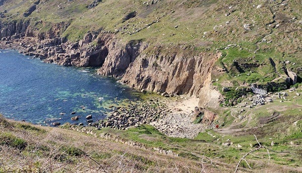 A photograph of a small coastal bay surrounded with cliffs, grassed at the top, with a small beach of sand and rocks giving way to a deep-blue sea.