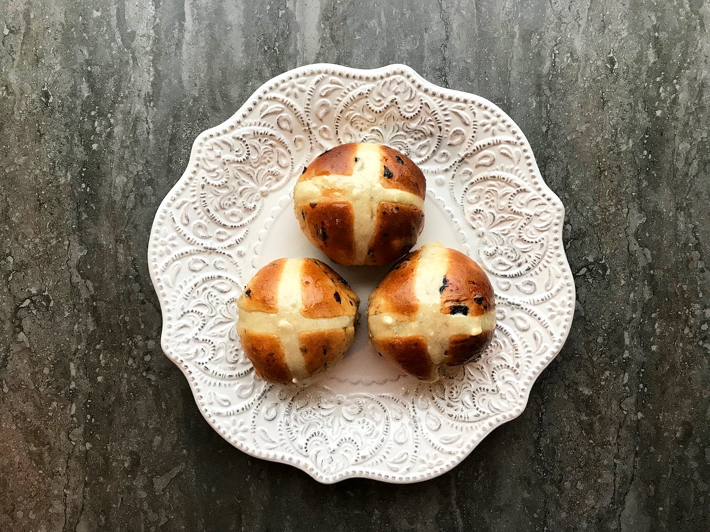 Three baked hot cross buns on an embossed white plate. The buns are a gorgeous umber, speckled with fruit. The paste cross is ivory-coloured in contrast.