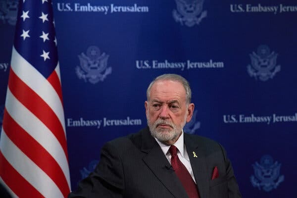 Mike Huckabee, the U.S. ambassador to Israel, in a dark suit and red tie. Behind him, a U.S. flag and a blue backdrop with "U.S. Embassy Jerusalem."
