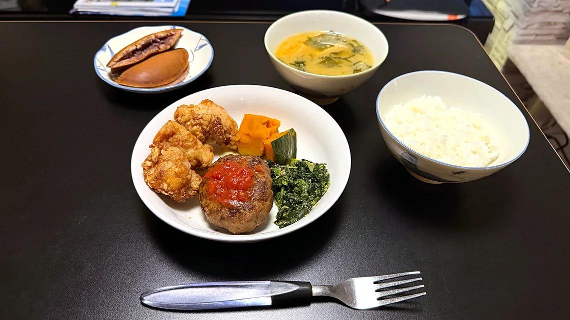 The matoil Set A dinner seen from above, with karaage, hamburger steak with tomato sauce, simmered pumpkin and sesame spinach on a white plate, miso soup with wakame, a bowl of white rice and two dorayaki