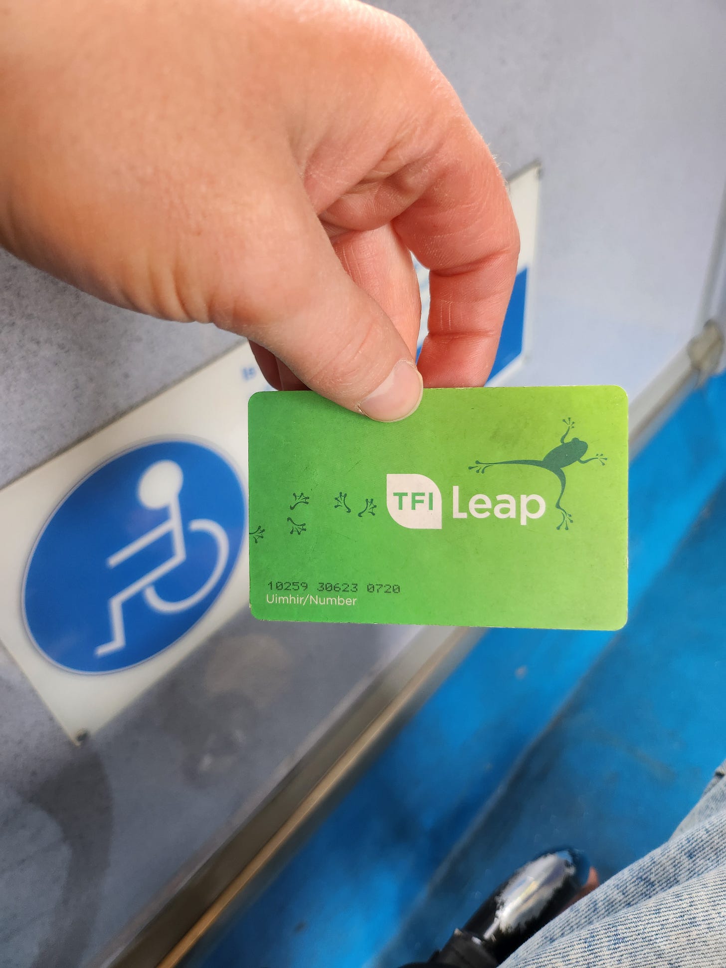 On Dublin Bus, I hold up a TFI Leap Card in my hand, just in front of the wheelchair symbol in the one space where wheelchair users can sit. 