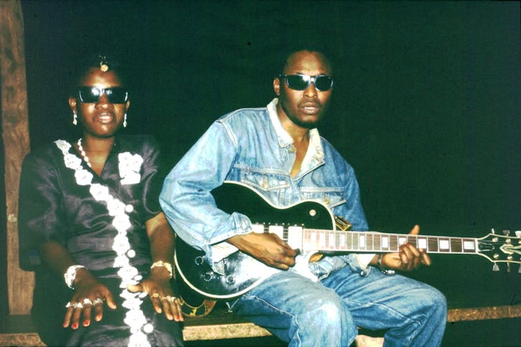 A young African couple in sunglasses, he in denim shirt and pants and she in a black and white dress, pose for a photo. He holds a guitar.