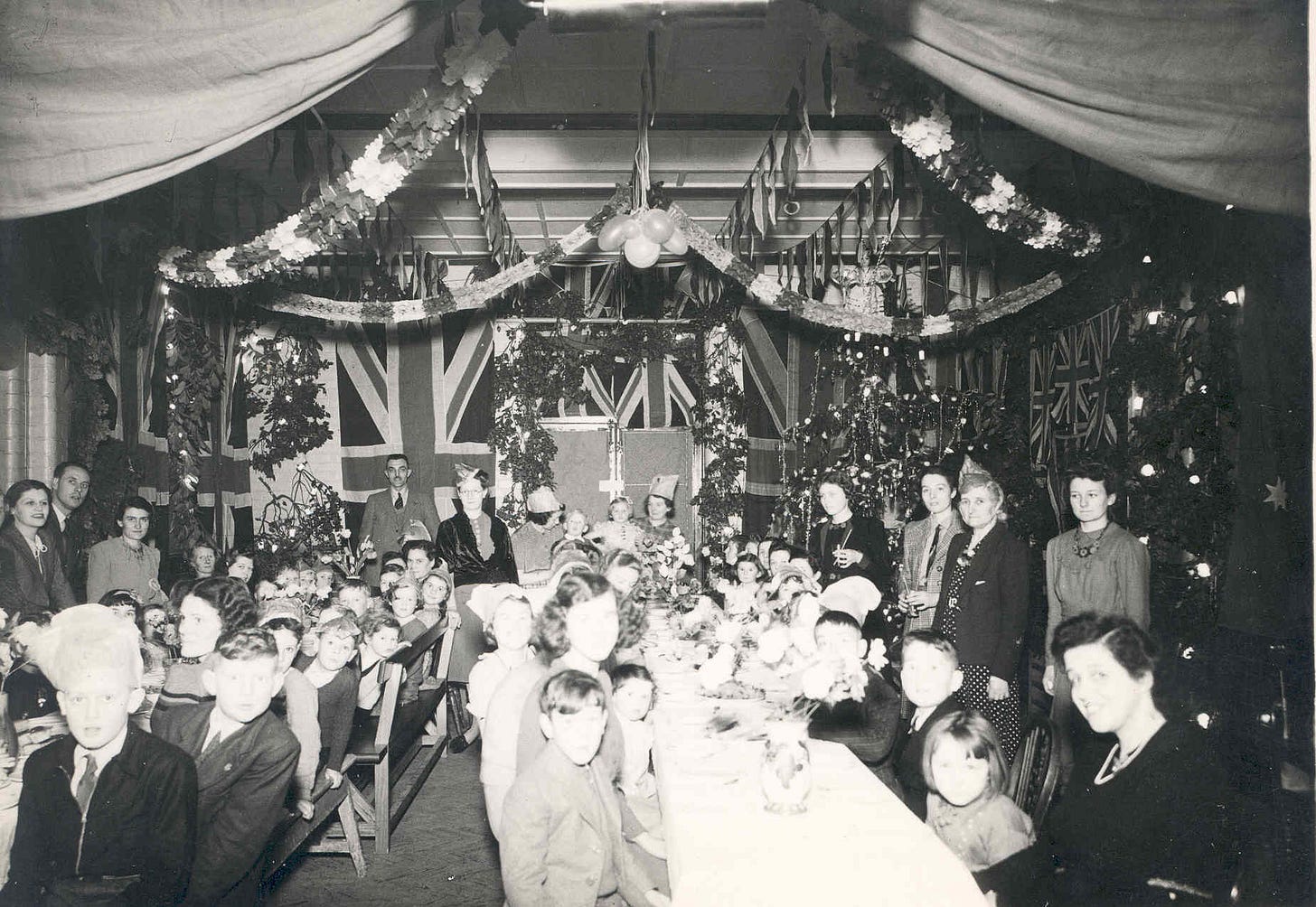 Children and adults sit at long tables in a hall decorated for christmas.