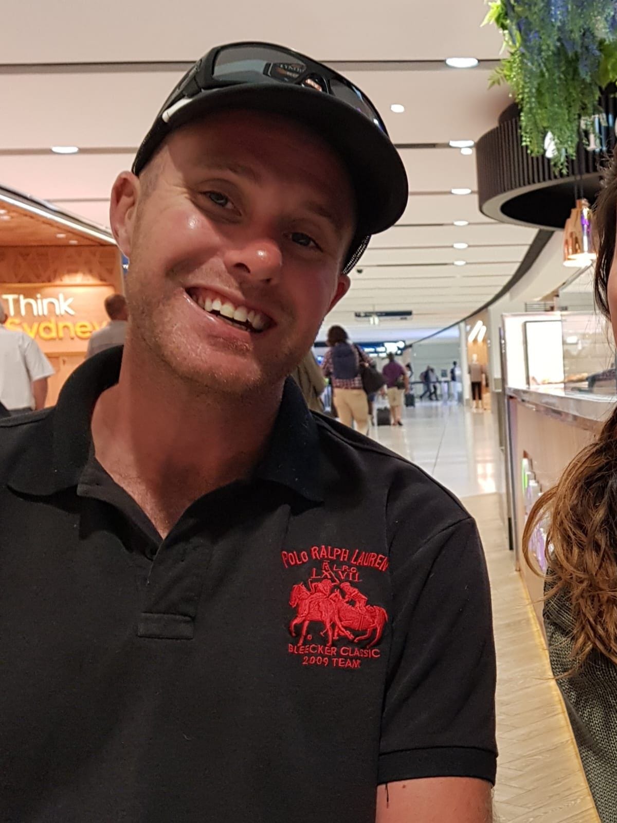 A smiling man in a black Polo Ralph Lauren shirt stands in a well-lit shopping mall with a casual and cheerful demeanor. The background features the text 'Think Sydney' and bustling shoppers. This image accompanies an article discussing Trump Coin, juxtaposing the lighthearted nature of collectibles with the weight of cultural and economic trends.