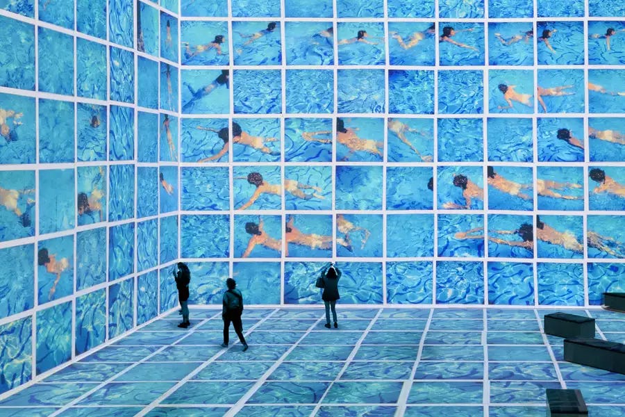 Three people stood in the David Hockney exhibition at Lightroom. His swimming pool paintings are shown on the screens. Three people stood in the David Hockney exhibition at Lightroom. His swimming pool paintings are shown on the screens.