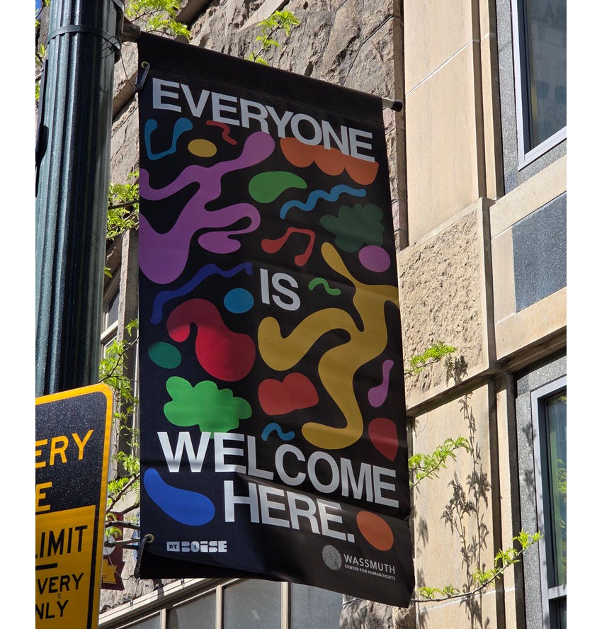 Lightpole banner reading 'Everyone is welcome here,' with colorful abstract blobs, musical notes, and two dancing human figures that resemble Keith Haring paintings if his figures were rounded off instead of angular.