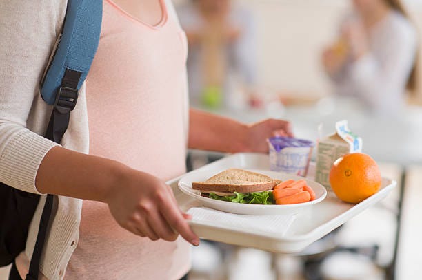 Student carrying a tray of lunch foods including a sandwich, baby carrots, orange, milk, and cereal cup