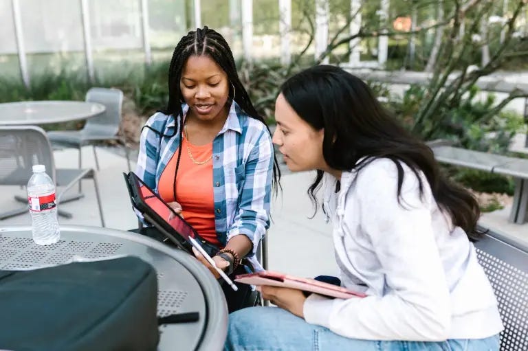 Two young women sit at an outdoor table, one holds a tablet, and both are looking at the tablet with interest. Two young women sit at an outdoor table, one holds a tablet, and both are looking at the tablet with interest.