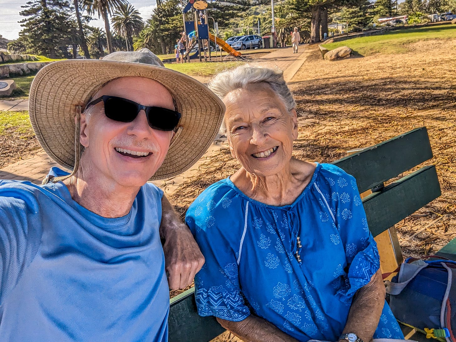 Michael wearing his sunhat and dark sunglasses sitting next to Colleen on a bench. 