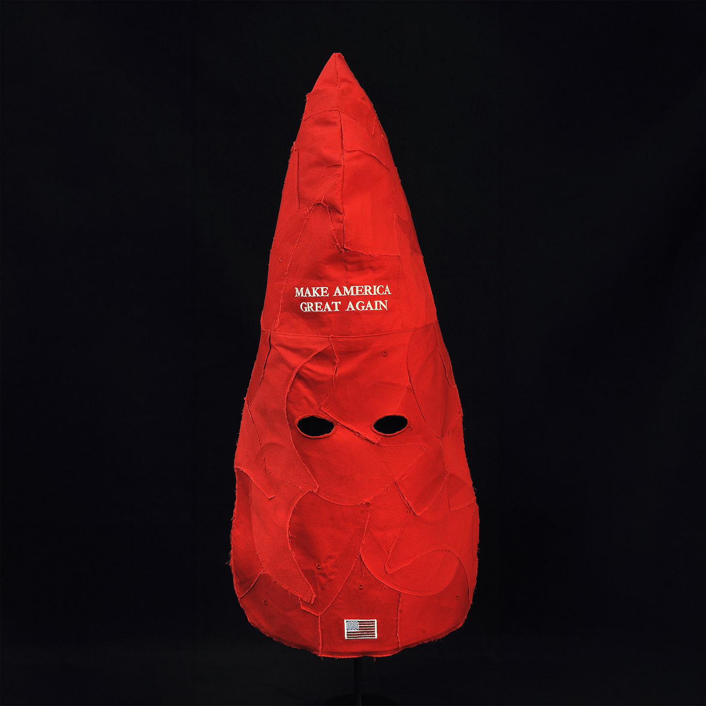 Image of The Hate Hat, an artwork by Kate Kretz. The piece is a freestanding Ku Klux Klan hood-shaped object, made entirely from deconstructed and sewn-together red MAGA hats. The words 'Make America Great Again' are prominently visible in white embroidery on the front. The object is set against a stark black background, emphasizing its bold form and color. Image of The Hate Hat, an artwork by Kate Kretz. The piece is a freestanding Ku Klux Klan hood-shaped object, made entirely from deconstructed and sewn-together red MAGA hats. The words 'Make America Great Again' are prominently visible in white embroidery on the front. The object is set against a stark black background, emphasizing its bold form and color.