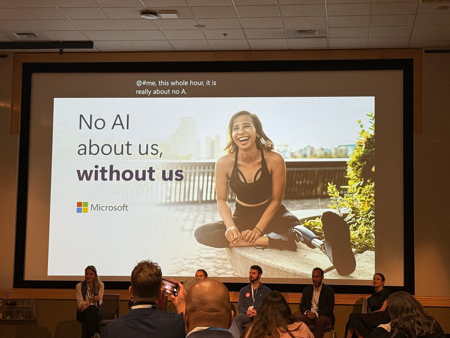 Projected Microsoft slide reading "No AI about us, without us," featuring a smiling woman with a prosthetic leg seated outdoors, with panelists and audience members visible below. Projected Microsoft slide reading "No AI about us, without us," featuring a smiling woman with a prosthetic leg seated outdoors, with panelists and audience members visible below.