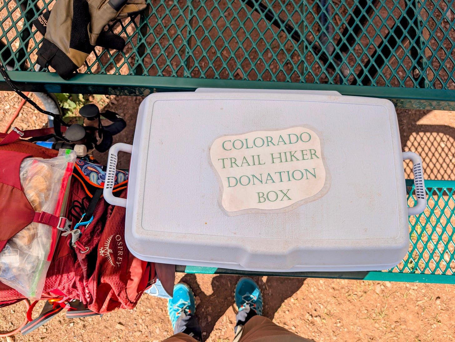 White cooler labeled Colorado Trail Hiker Donation Box on a green metal picnic table, with a backpack and trekking poles nearby. White cooler labeled Colorado Trail Hiker Donation Box on a green metal picnic table, with a backpack and trekking poles nearby.