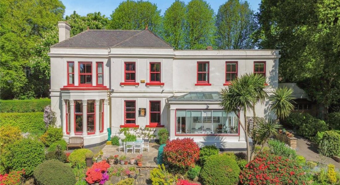 A house exterior with white walls and red windows, and trees and greenery around it