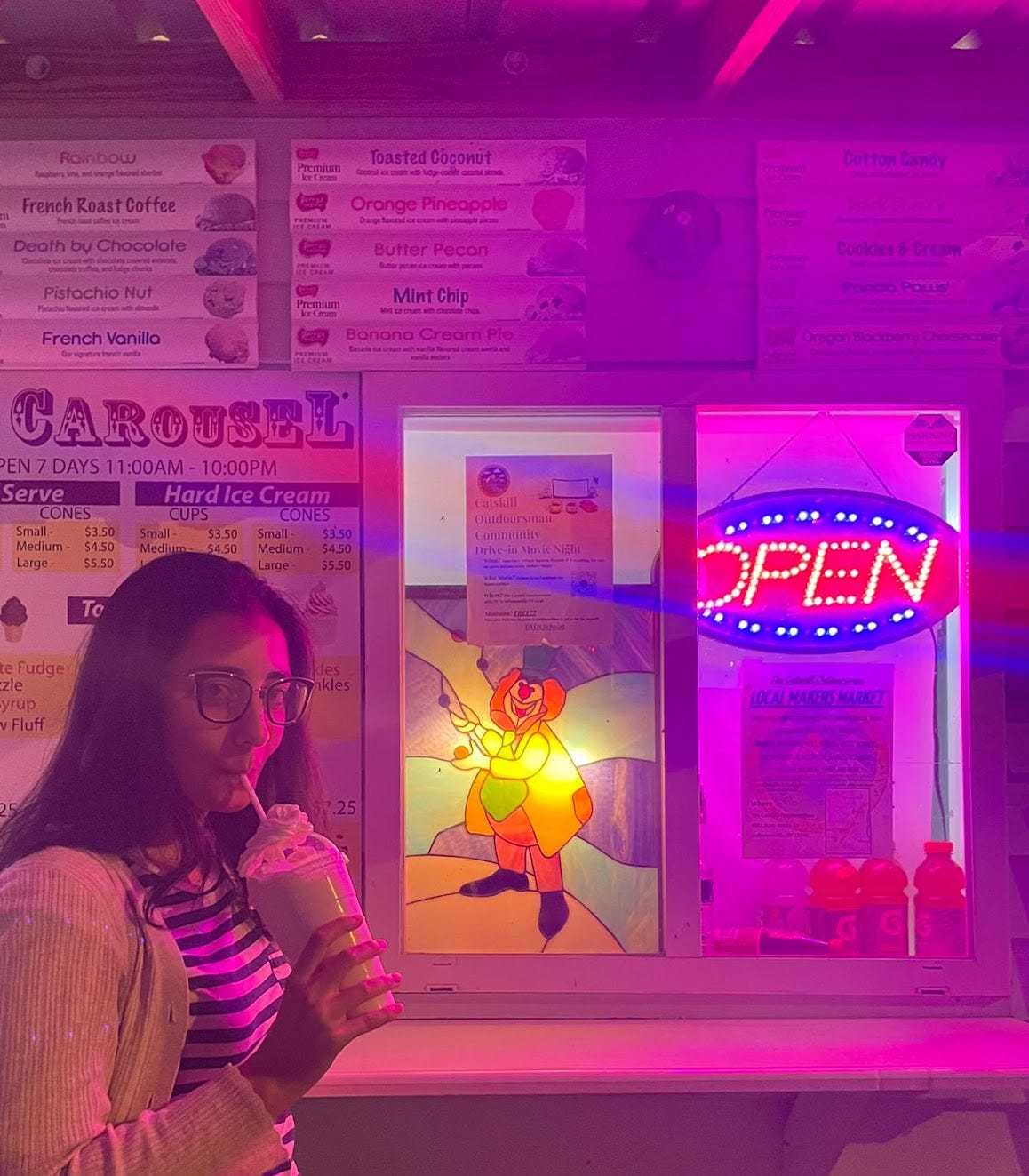 A young woman with black hair and glasses drinks a milkshake in front of an ice cream stand A young woman with black hair and glasses drinks a milkshake in front of an ice cream stand