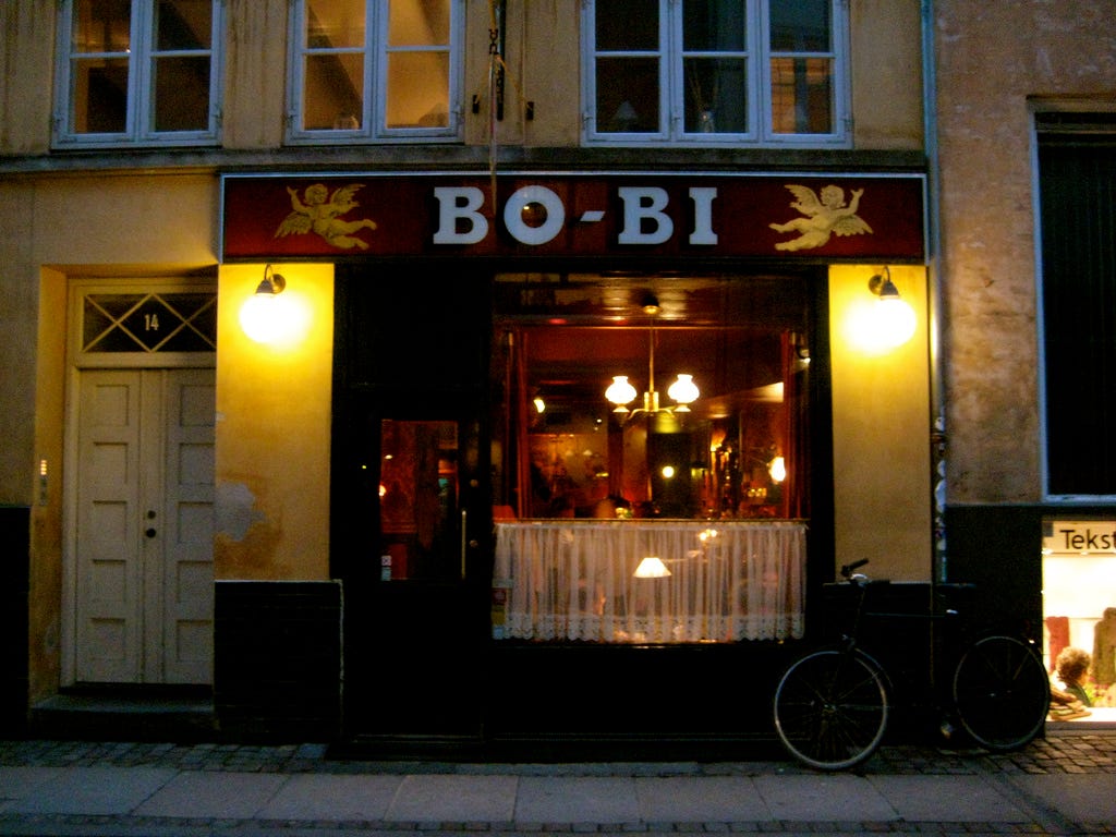Bo-Bi bar at dusk, cafe curtain in the street front window with warm reddish glow inside - unsurprisingly a bike is parked outside