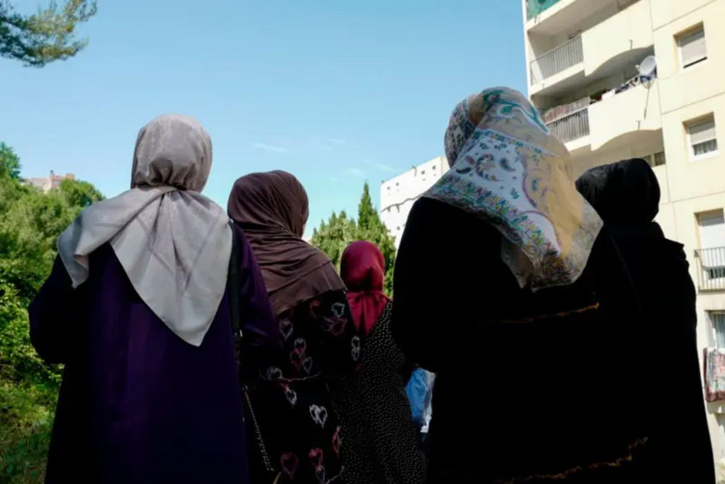 A group of five women in headscarves with their backs to the camera stans in an urban area with greenery on the left side and a multi-storey residential building on the right