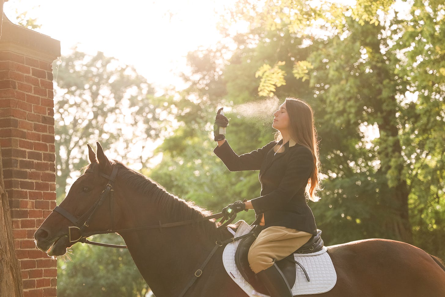 A woman in equestrian clothing sits upright on a horse, holding a spray bottle and spraying a fine mist on her face. The sunlight streams through the trees, casting a warm glow over the scene, and a brick column is visible to the left.