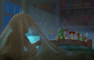 In a scene from Disney and Pixar’s Toy Story 5, we are looking at a darkened bedroom from the head of a twin bed. Directly in front of us, on the left side of the image, a mysterious person, probably a child, is silhouetted beneath a blue polka-dot sheet, holding a computer tablet that has them entranced. At the end of the bed, peeking over the foot of the bed and lit by a beam of moonlight, are many of the Toy Story characters, including Woody, Jessie, and Buzz.