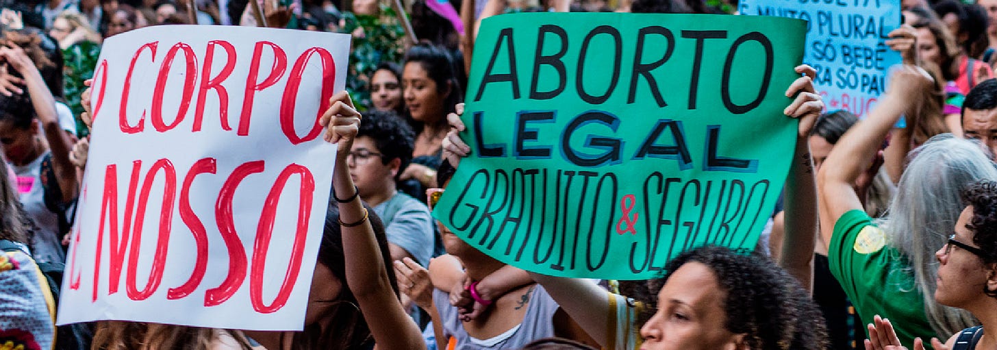 Una foto orizzontale di una protesta per l'aborto. I manifestanti tengono in mano cartelli con slogan a favore della legalizzazione dell'aborto. Al centro dell'immagine, una donna vestita di nero con una mascherina bianca alza un pugno. Sullo sfondo, si vedono altri manifestanti e striscioni con scritte in portoghese.