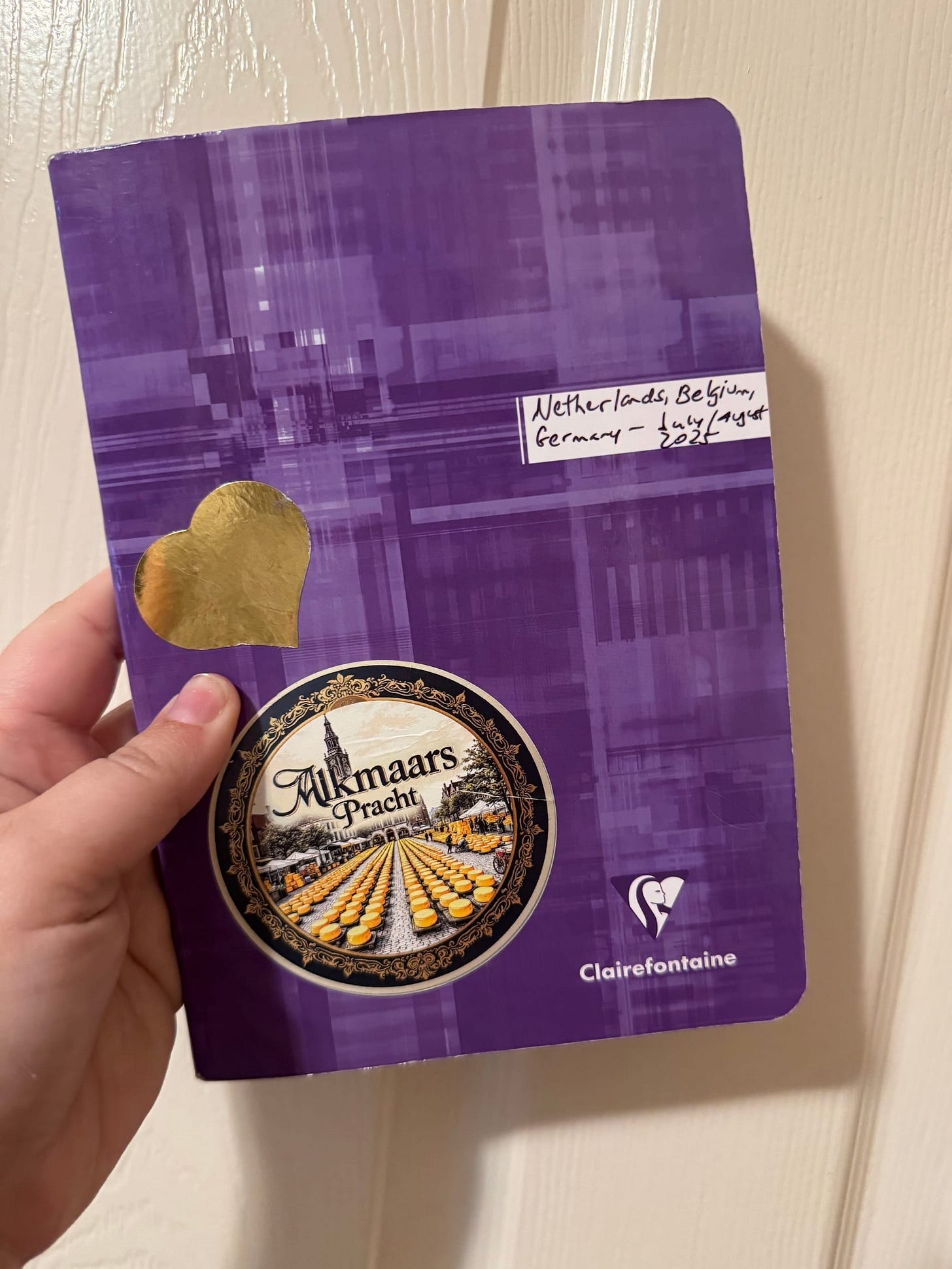 A light-skinned hand holds up a purple notebook with stickers and some wear on it in front of an off-white door