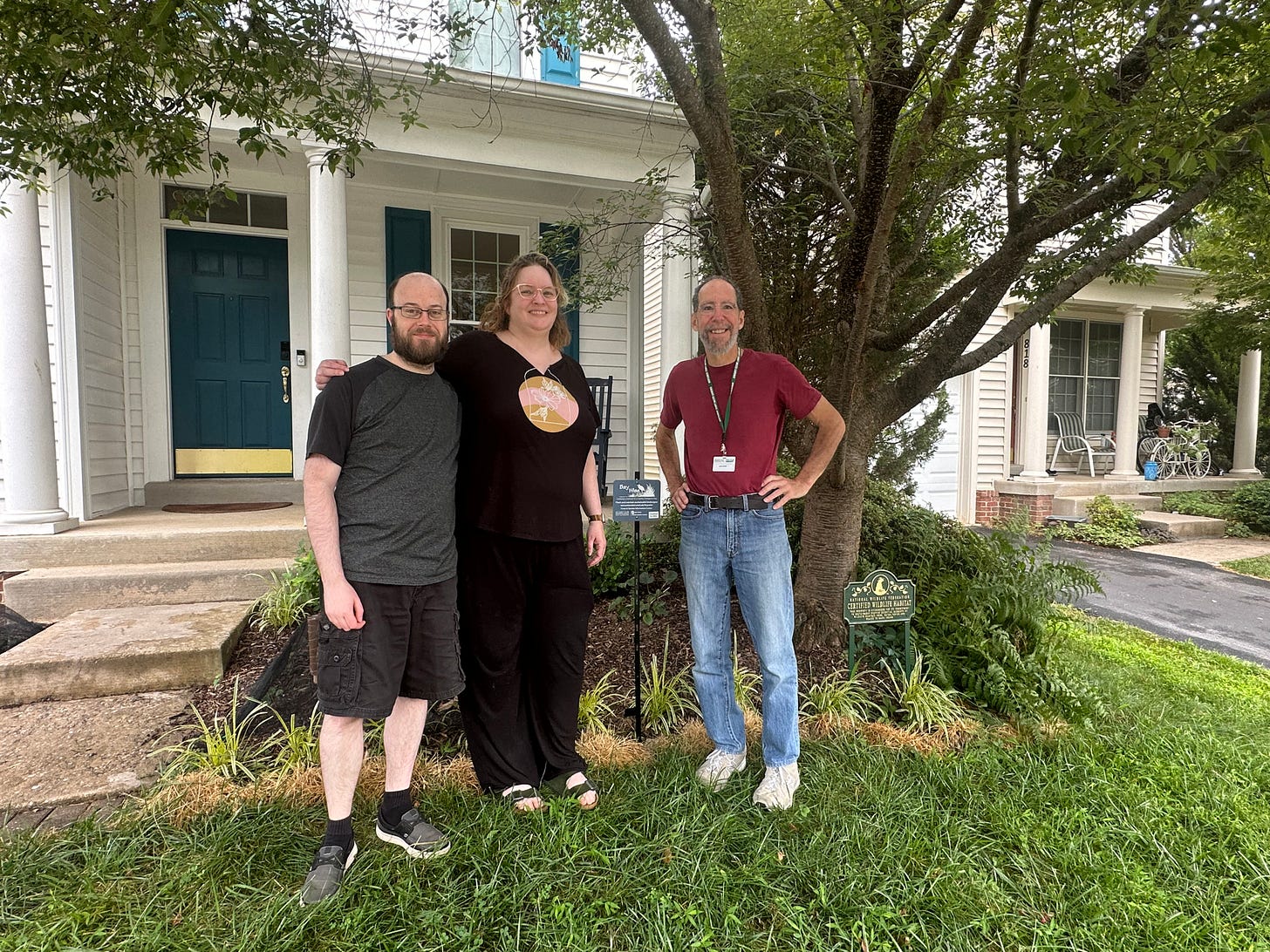 Three people are standing in front of a white house with a teal door and blue shutters. The person on the left is a bearded man wearing glasses, a dark gray t-shirt, and black shorts. The person in the middle is a woman with blonde hair, wearing glasses, a black t-shirt with a circular design, and black pants. The person on the right is a man with a beard, wearing a red t-shirt, blue jeans, and a green lanyard with a name tag. They are standing on a well-maintained lawn with a tree and various plants. There is a green Bay-Wise certification sign near the tree, indicating that the property is certified as a sustainable landscape and NWF wildlife habitat. Three people are standing in front of a white house with a teal door and blue shutters. The person on the left is a bearded man wearing glasses, a dark gray t-shirt, and black shorts. The person in the middle is a woman with blonde hair, wearing glasses, a black t-shirt with a circular design, and black pants. The person on the right is a man with a beard, wearing a red t-shirt, blue jeans, and a green lanyard with a name tag. They are standing on a well-maintained lawn with a tree and various plants. There is a green Bay-Wise certification sign near the tree, indicating that the property is certified as a sustainable landscape and NWF wildlife habitat.