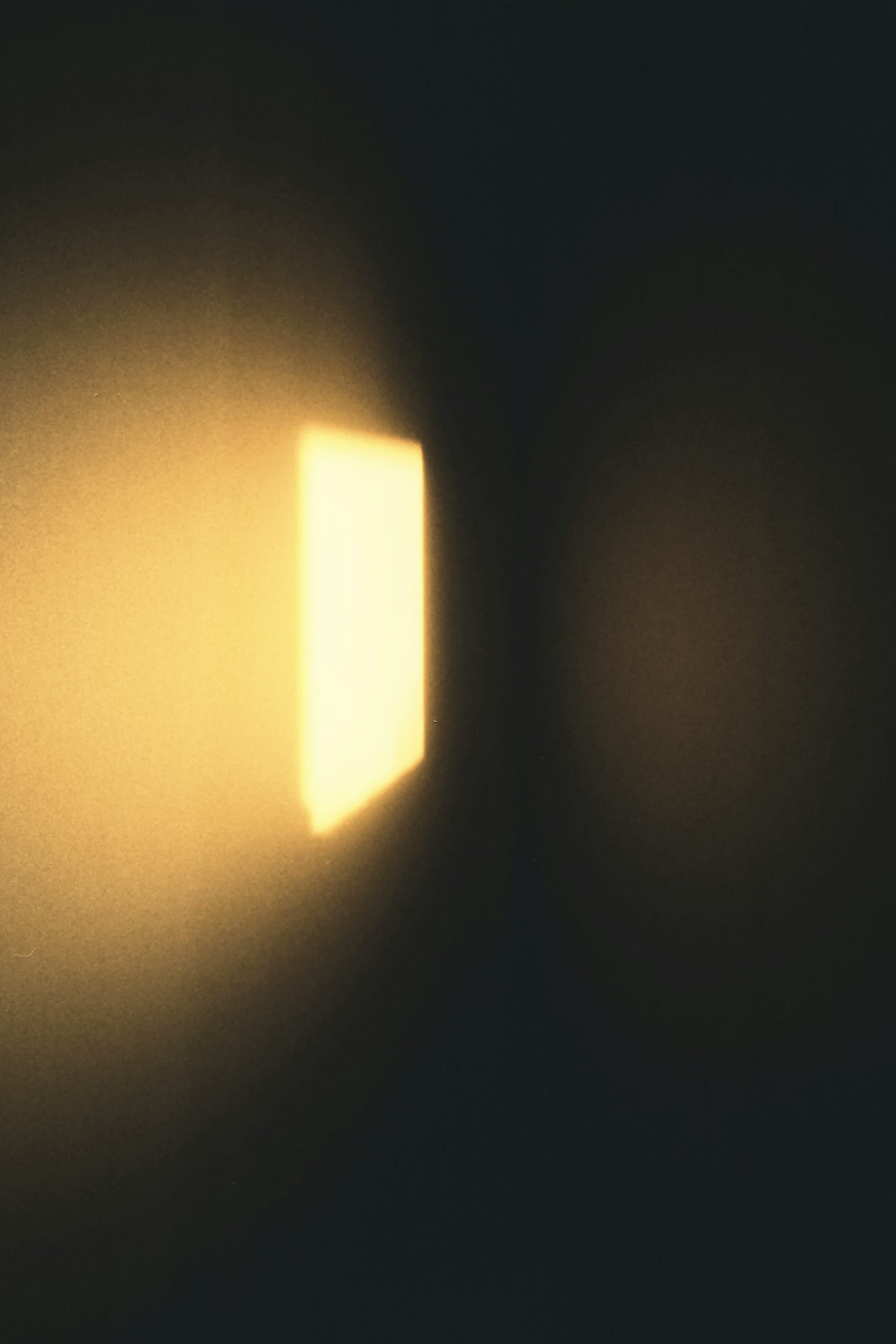 A soft, golden rectangle of light falls across a dark wall, creating a blurred, glowing window-shaped patch that fades into deep shadow. The scene feels quiet, minimal, and atmospheric—as if sunlight is slipping into an otherwise dim room.