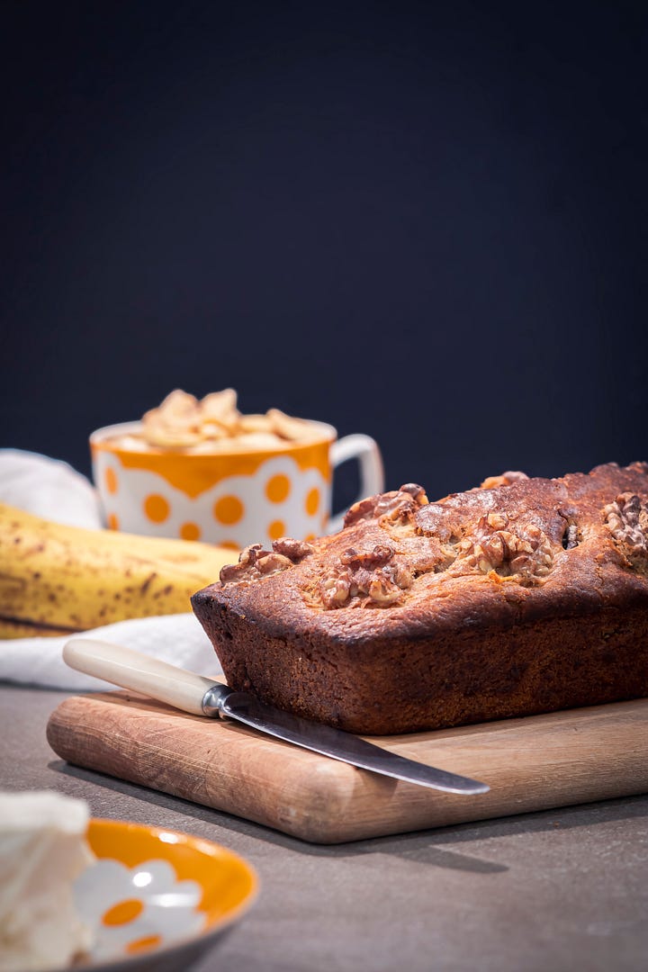 Homemade banana bread topped with pecans, shown whole and sliced, topped with scoops of snow-ball imitating cream cheese. 